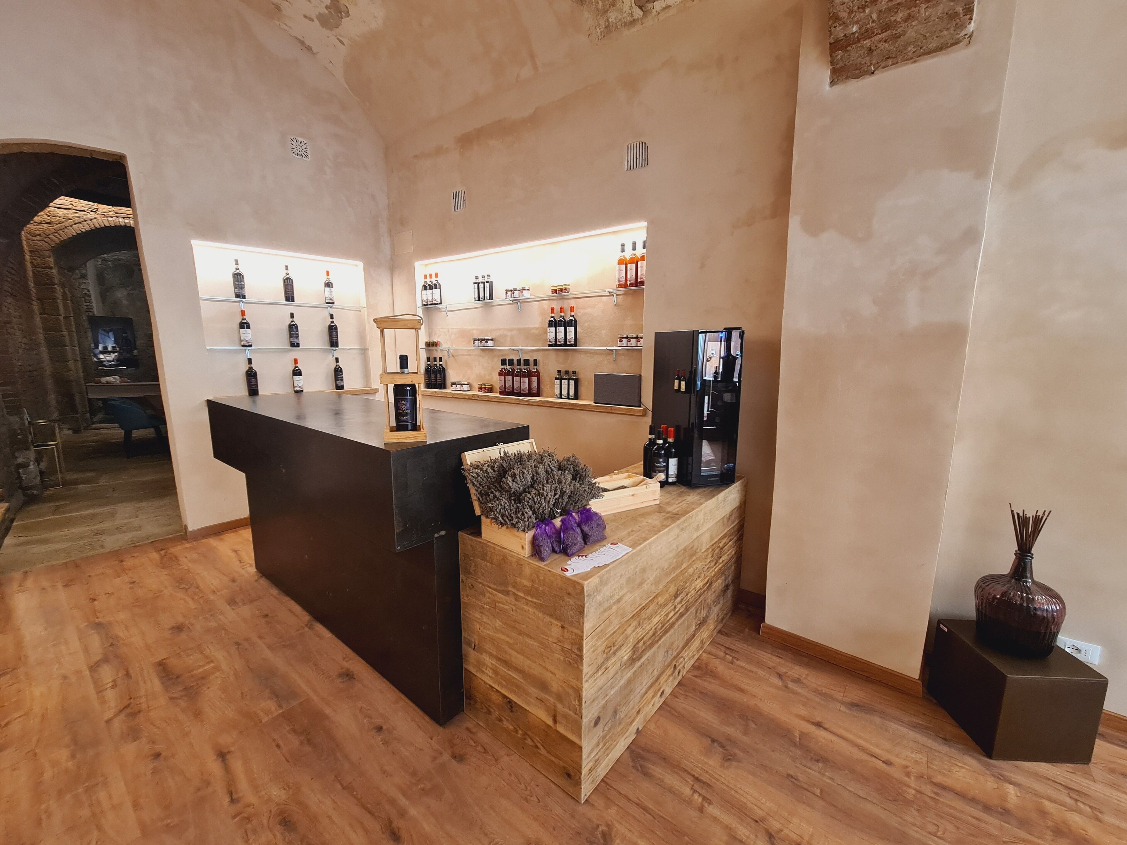 Un espace d’accueil avec un comptoir en bois, des étagères remplies de boissons en bouteille, un réfrigérateur noir et un vase décoratif.