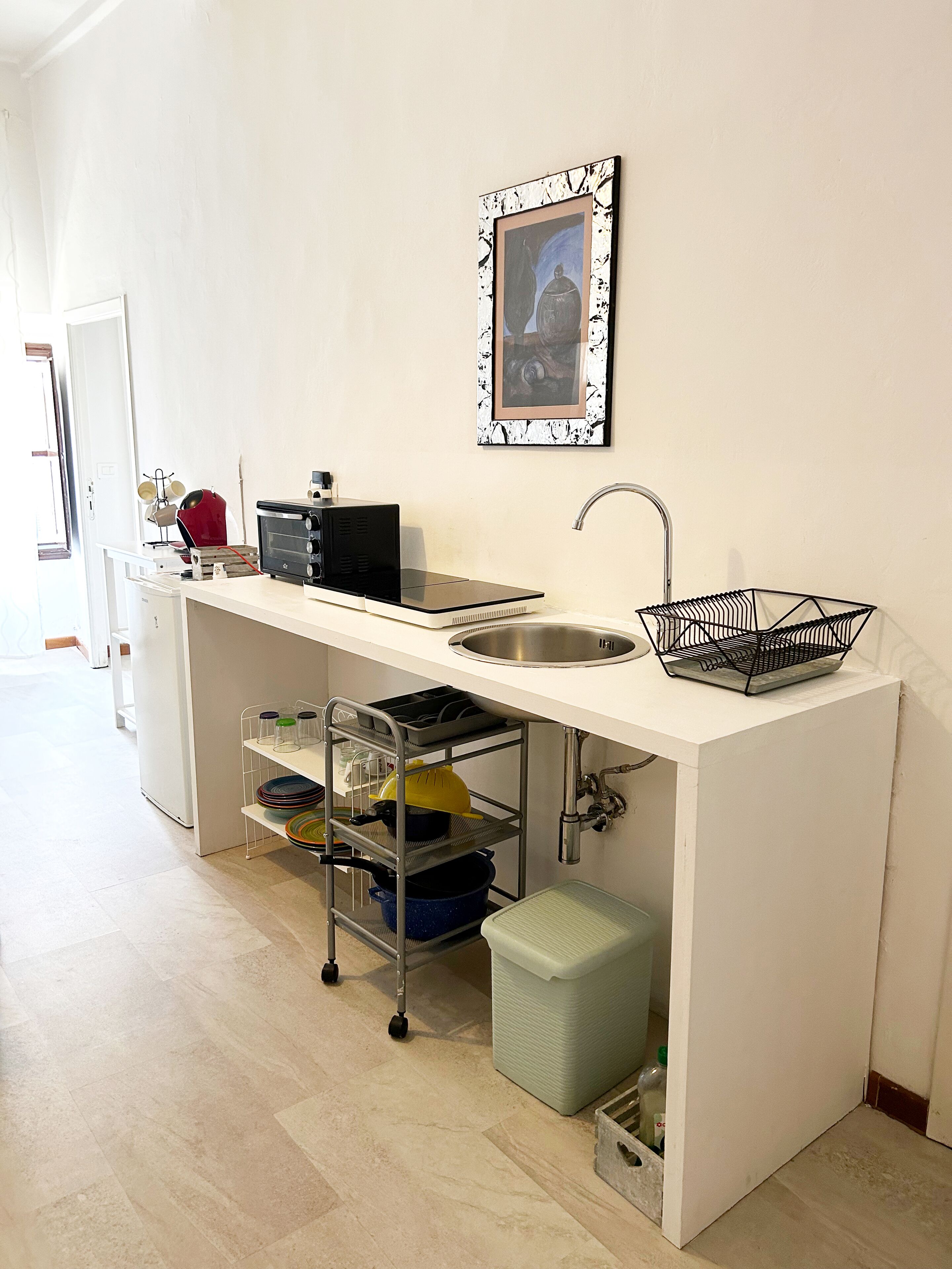 Una cocina compacta con pileta integrada, un microondas y una pequeña mesada. Se observa una rejilla metálica con vajilla y un bote de basura debajo.