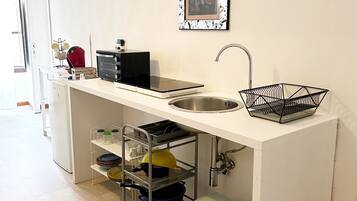 Una cocina compacta con pileta integrada, un microondas y una pequeña mesada. Se observa una rejilla metálica con vajilla y un bote de basura debajo.