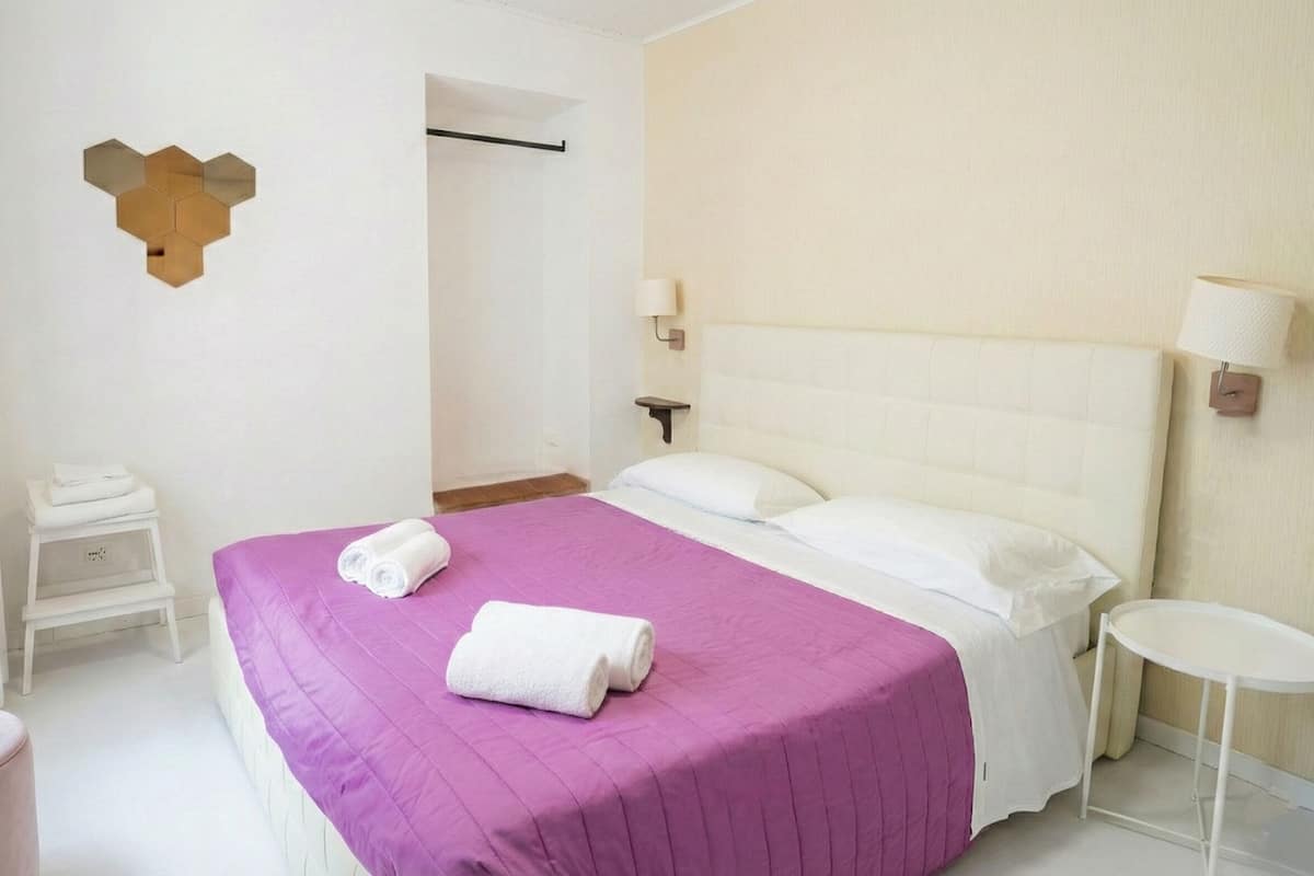 A hotel room with a bed, a bedside table, a small stool, a wall-mounted light, and a wall decoration.