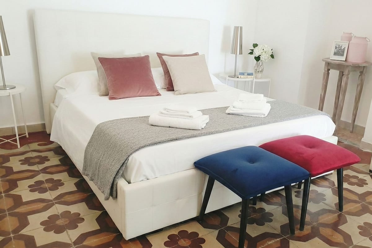 A neatly made bed with pillows, a bedside table with a lamp, a small table with a vase and picture, and a bench with cushions.