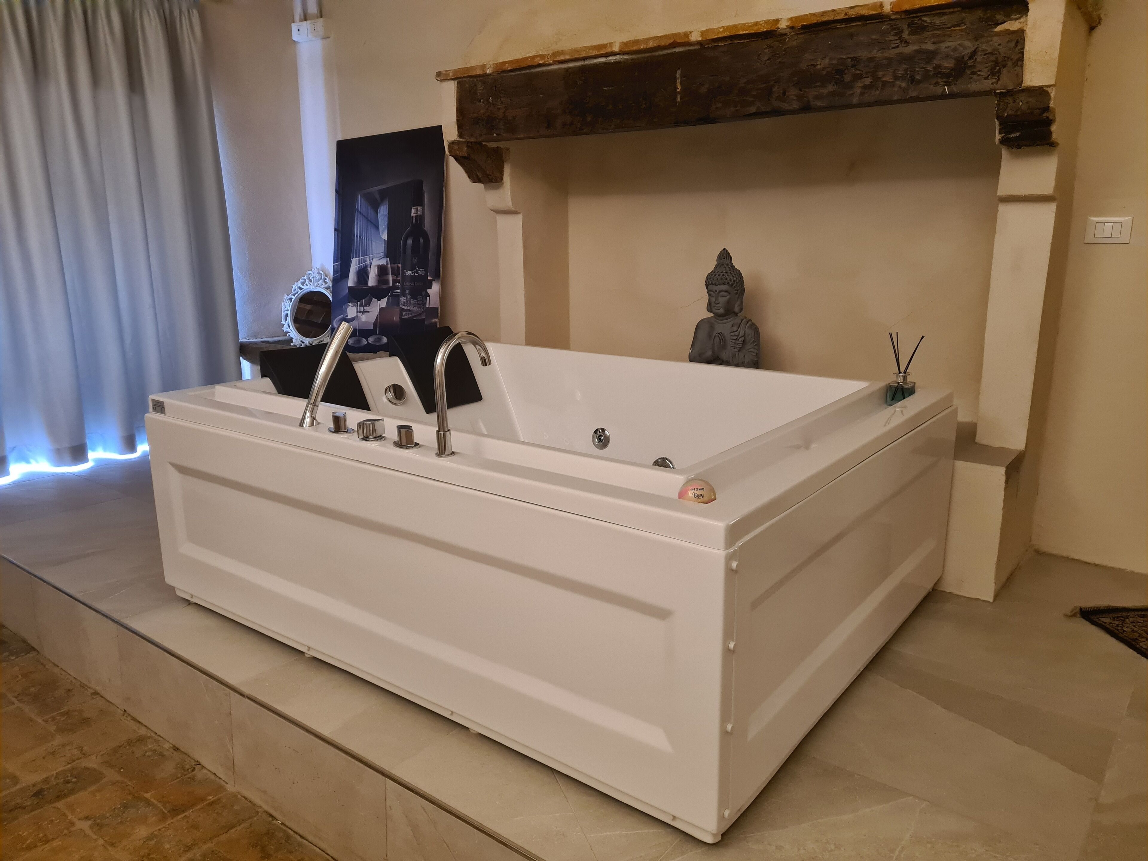 Una bañera blanca independiente con dos grifos y un desagüe, ubicada en un cuarto con piso de baldosas y una viga de madera en el techo.