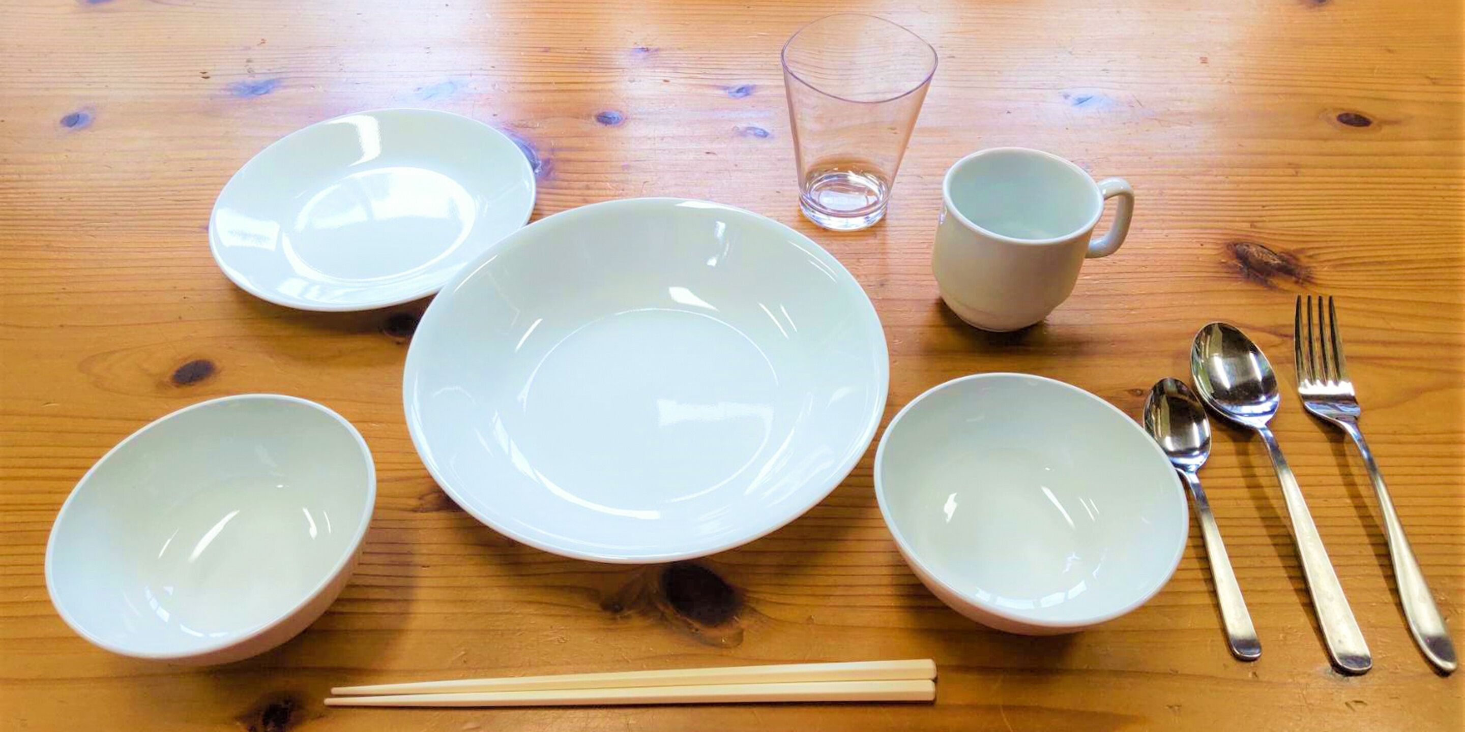 A wooden table with five white bowls, a glass, a white mug, and a set of silverware.