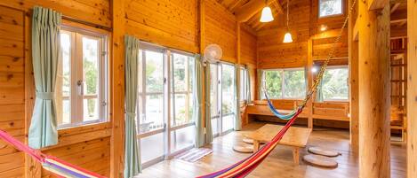 A wooden cabin interior with large windows, a hammock, and a wooden bench.