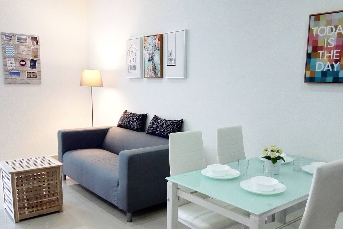 A modern living room with a grey sofa, a glass dining table set for two, and a small side table.