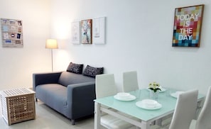 Una sala moderna con un sofá gris, una mesa de comedor de vidrio para dos comensales y una mesita auxiliar.