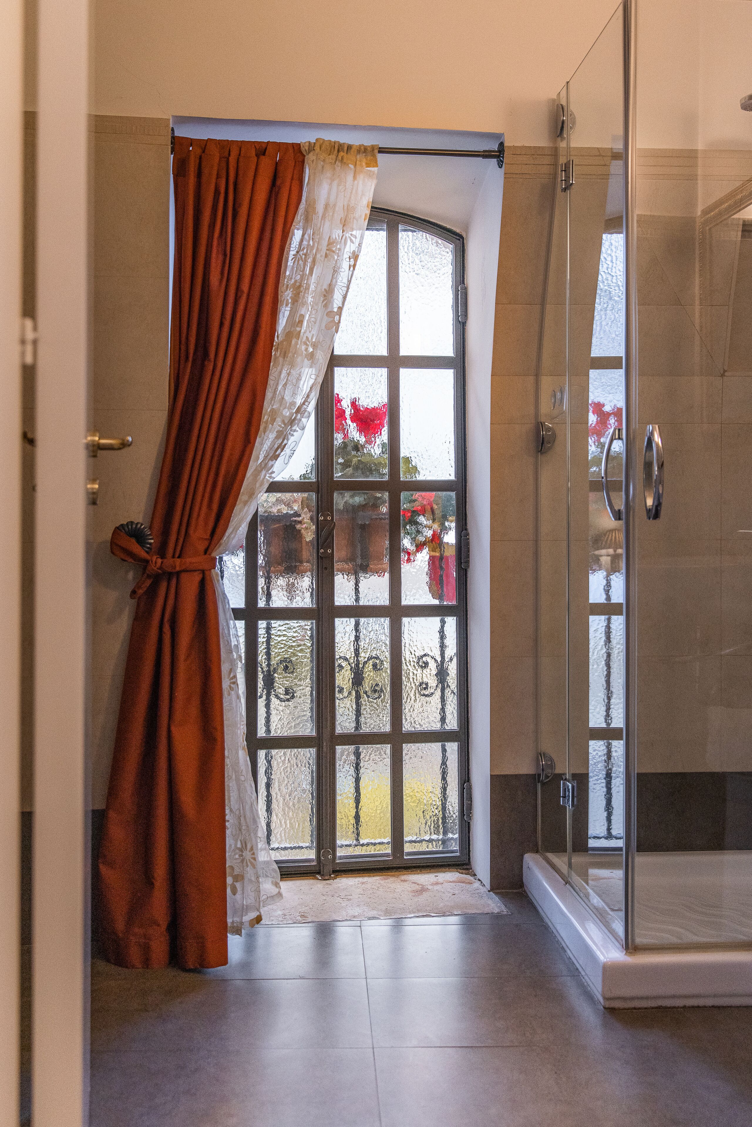 A bathroom with a glass shower enclosure, a large window with a view of a garden, and a door with a curtain.