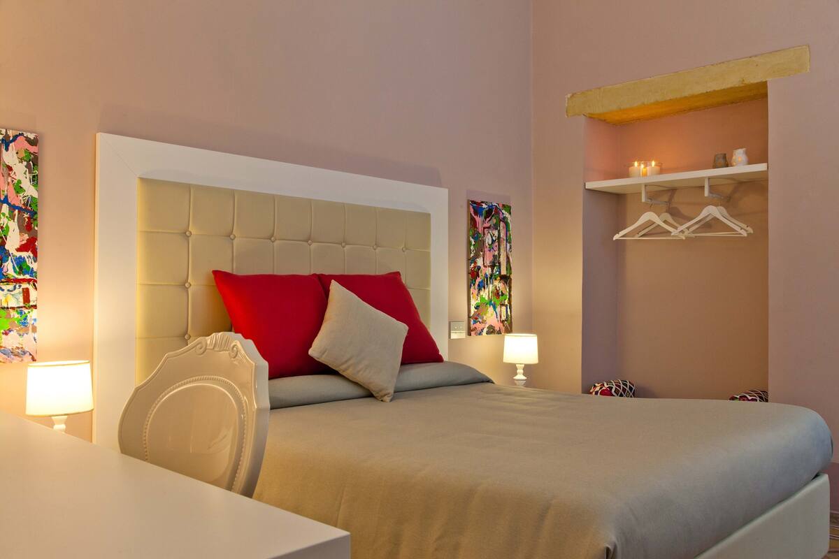 Une chambre d’hôtel avec un lit, des tables de chevet, une chaise, une lampe suspendue, une étagère avec des vêtements et un tableau au mur.