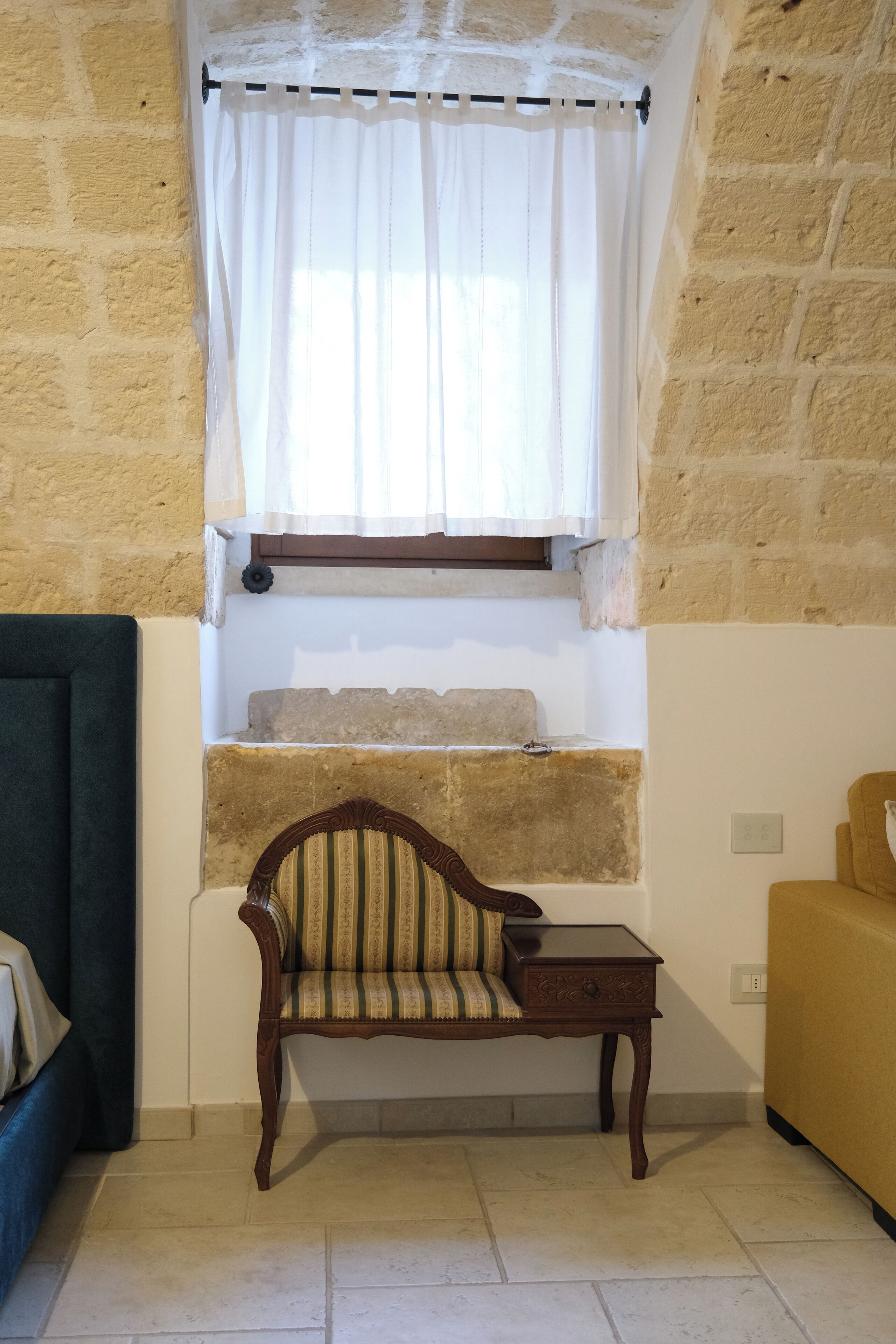 Una habitación con pared de piedra, un banco de madera con cojín, una mesita y una ventana con cortina blanca.