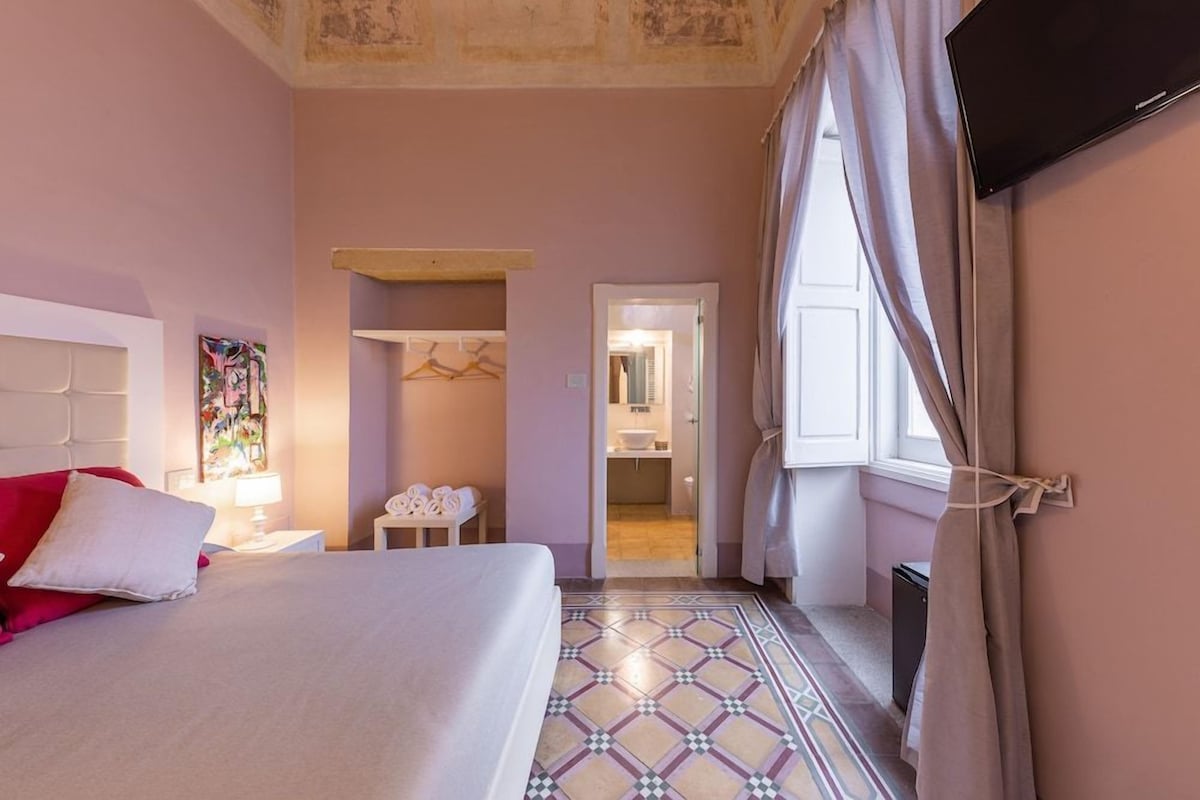 Habitación de hotel con cama, mesita de noche, lámpara, perchero y baño visible a través de una puerta abierta.