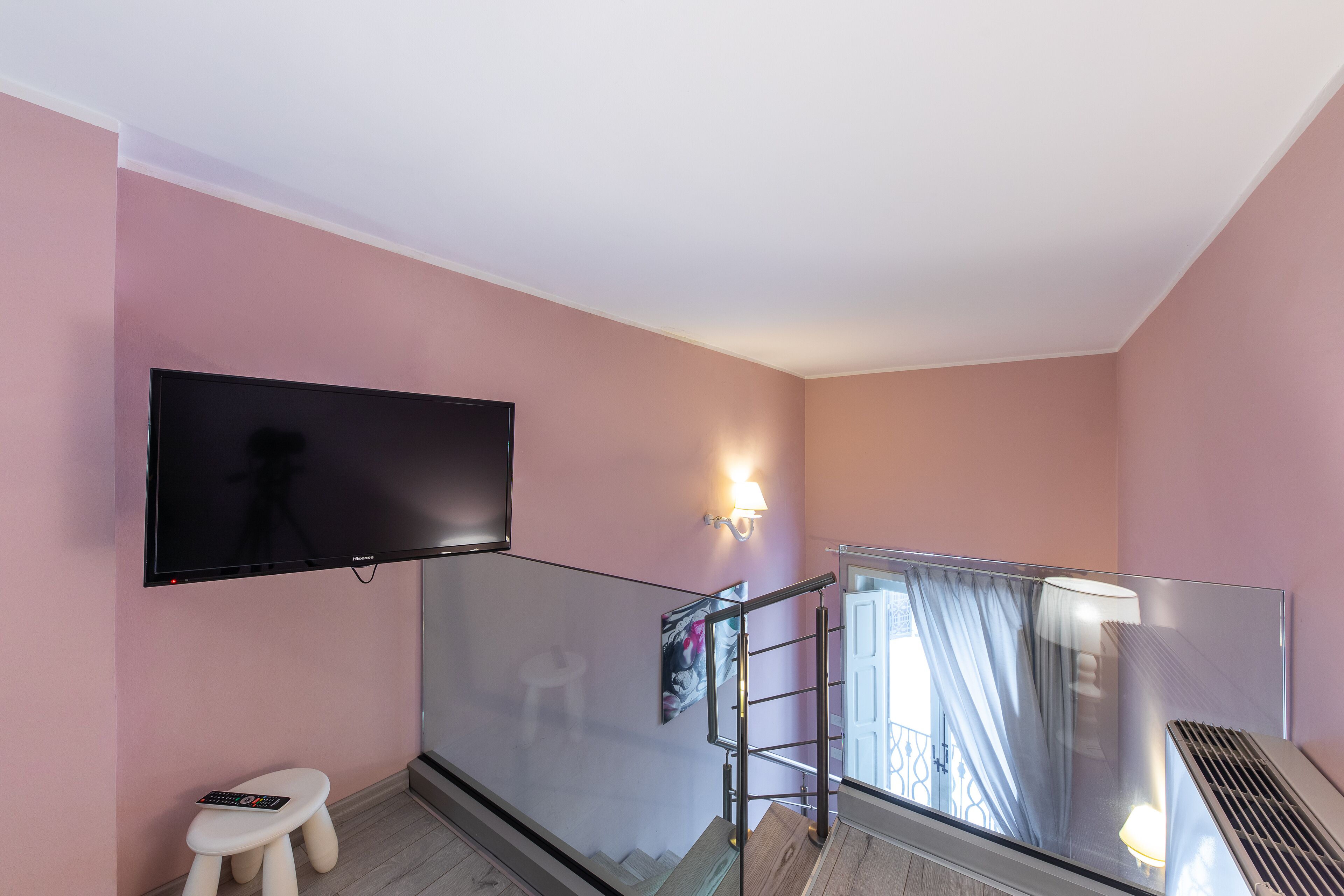 Habitación de hotel moderna con un televisor de pantalla plana grande montado en la pared, un taburete pequeño, barandilla de cristal y una ventana con cortinas.