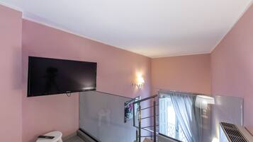 Habitación de hotel moderna con un televisor de pantalla plana grande montado en la pared, un taburete pequeño, barandilla de cristal y una ventana con cortinas.