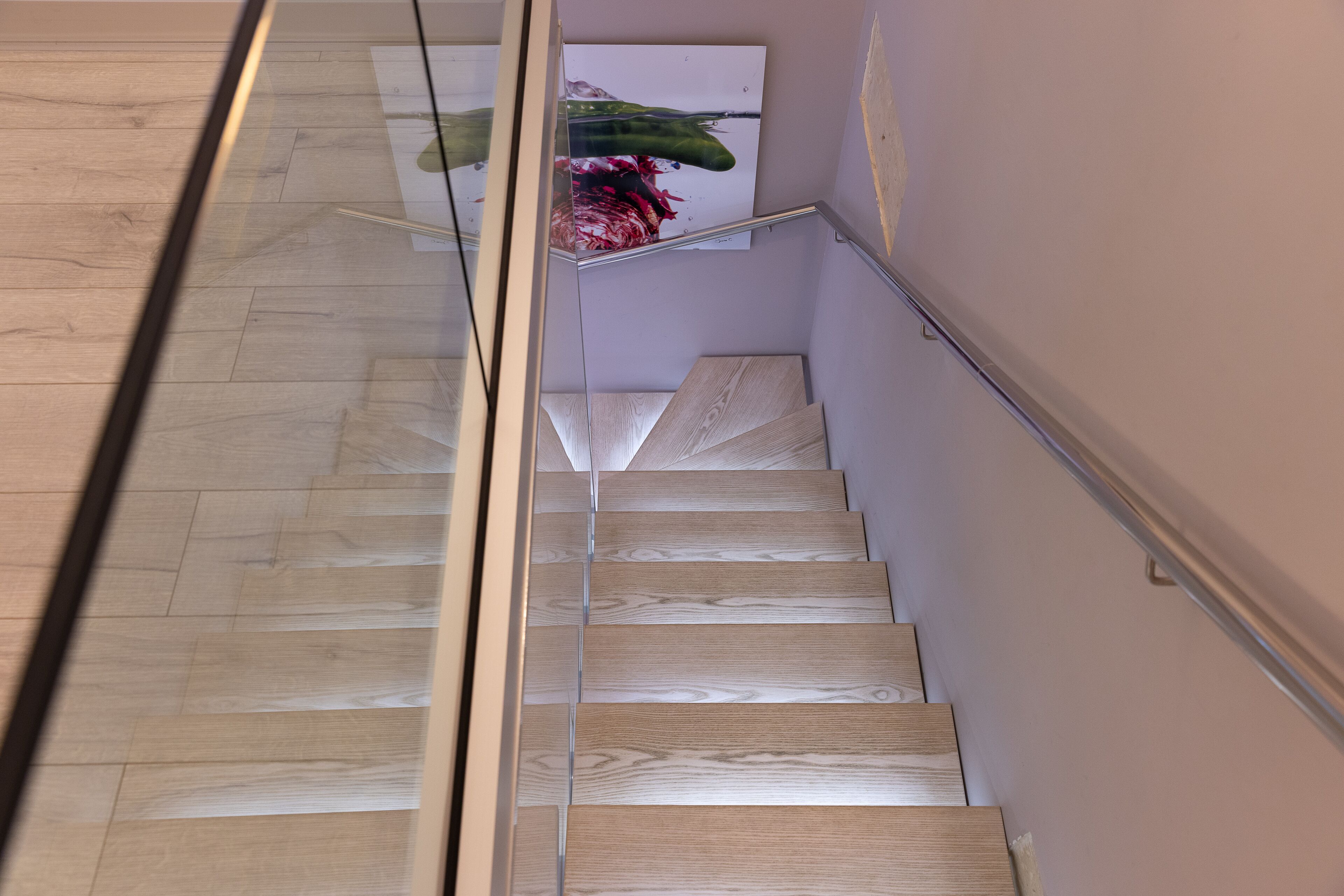Eine Holztreppe mit Glastrennstäben, die zu einer Zwischenebene mit einem gerahmten Kunstwerk führt.