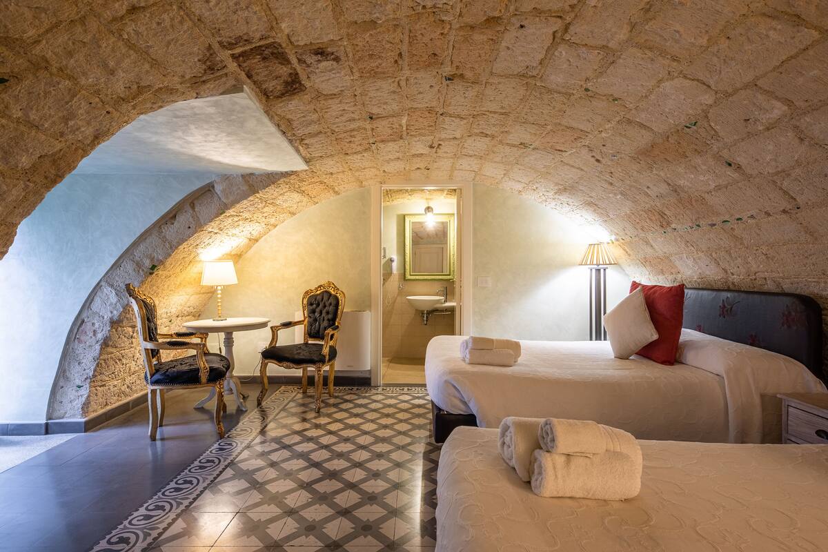 Une pièce chaleureuse avec un plafond en pierre, deux lits, une chaise, une petite table et une salle de bain visible à travers une arche.