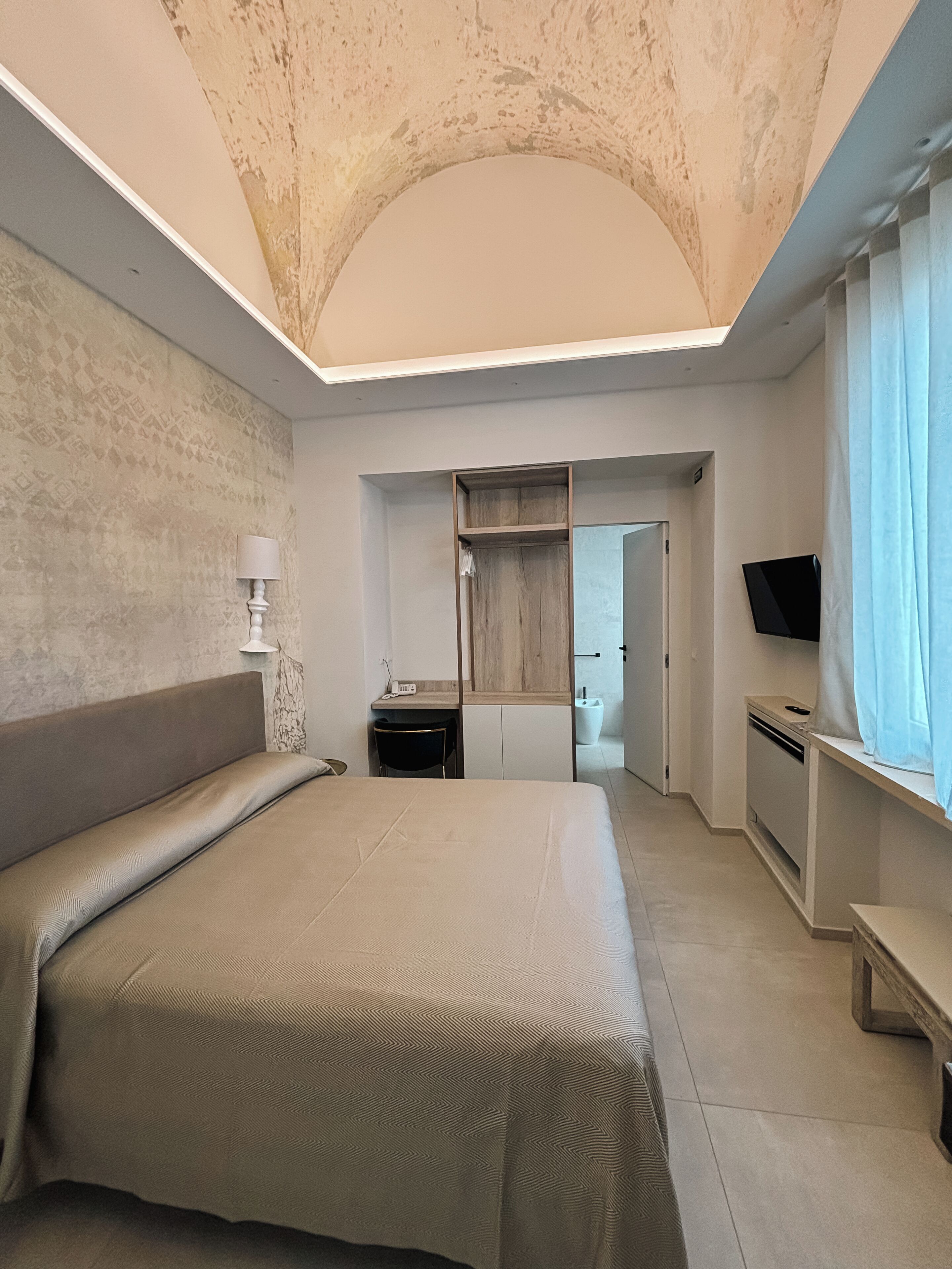 A bedroom with a large bed, a desk, a television, and a bathroom visible through an open door.