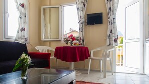 Una habitación con sofá, una mesa de centro de vidrio con un jarrón, una mesa roja con sillas, un televisor en la pared y un balcón con vistas.