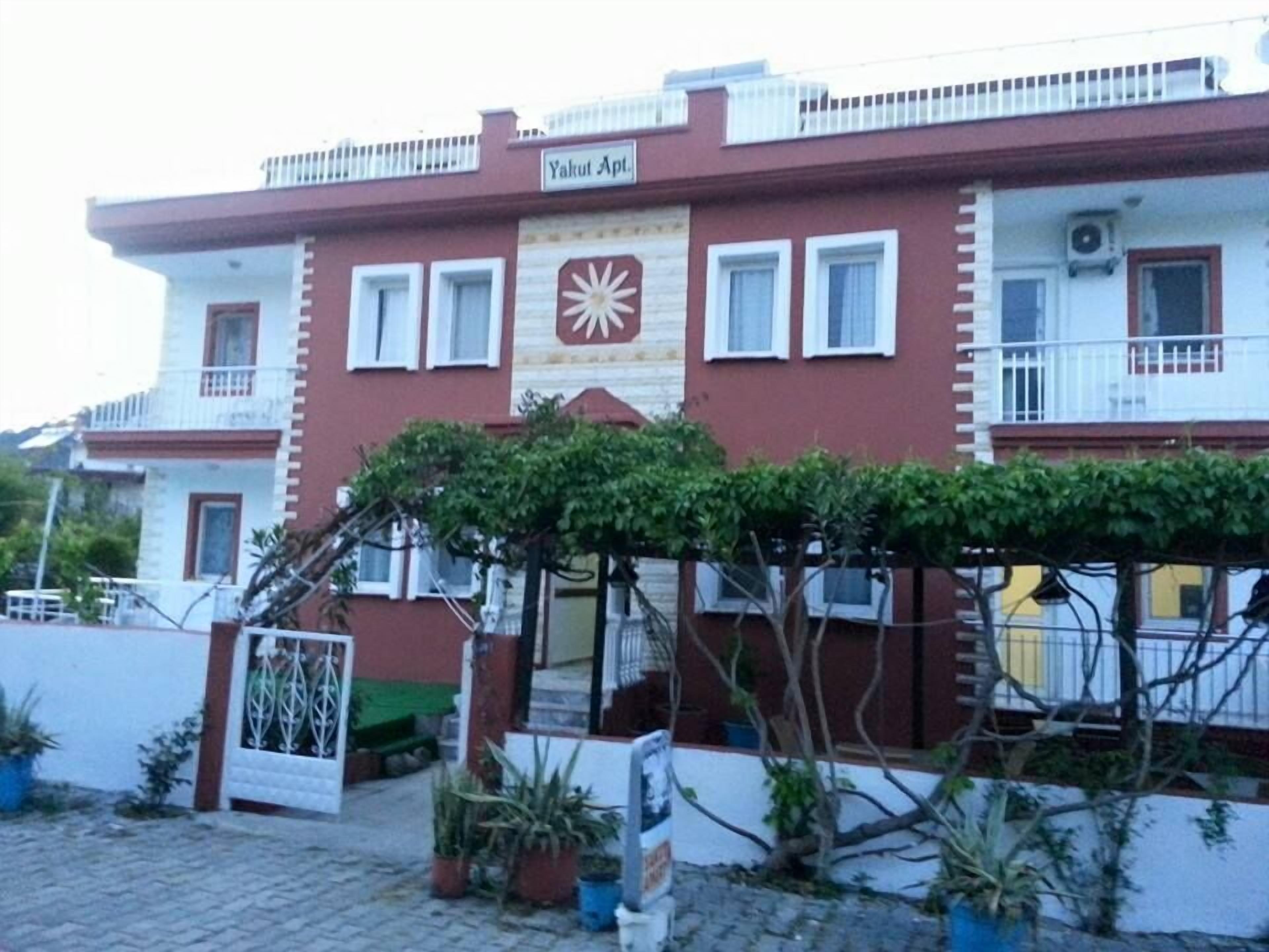 Ein zweistöckiges Gebäude mit roter und weißer Fassade, einem Balkon im zweiten Stock und einem kleinen Garten mit Blumentöpfen davor.