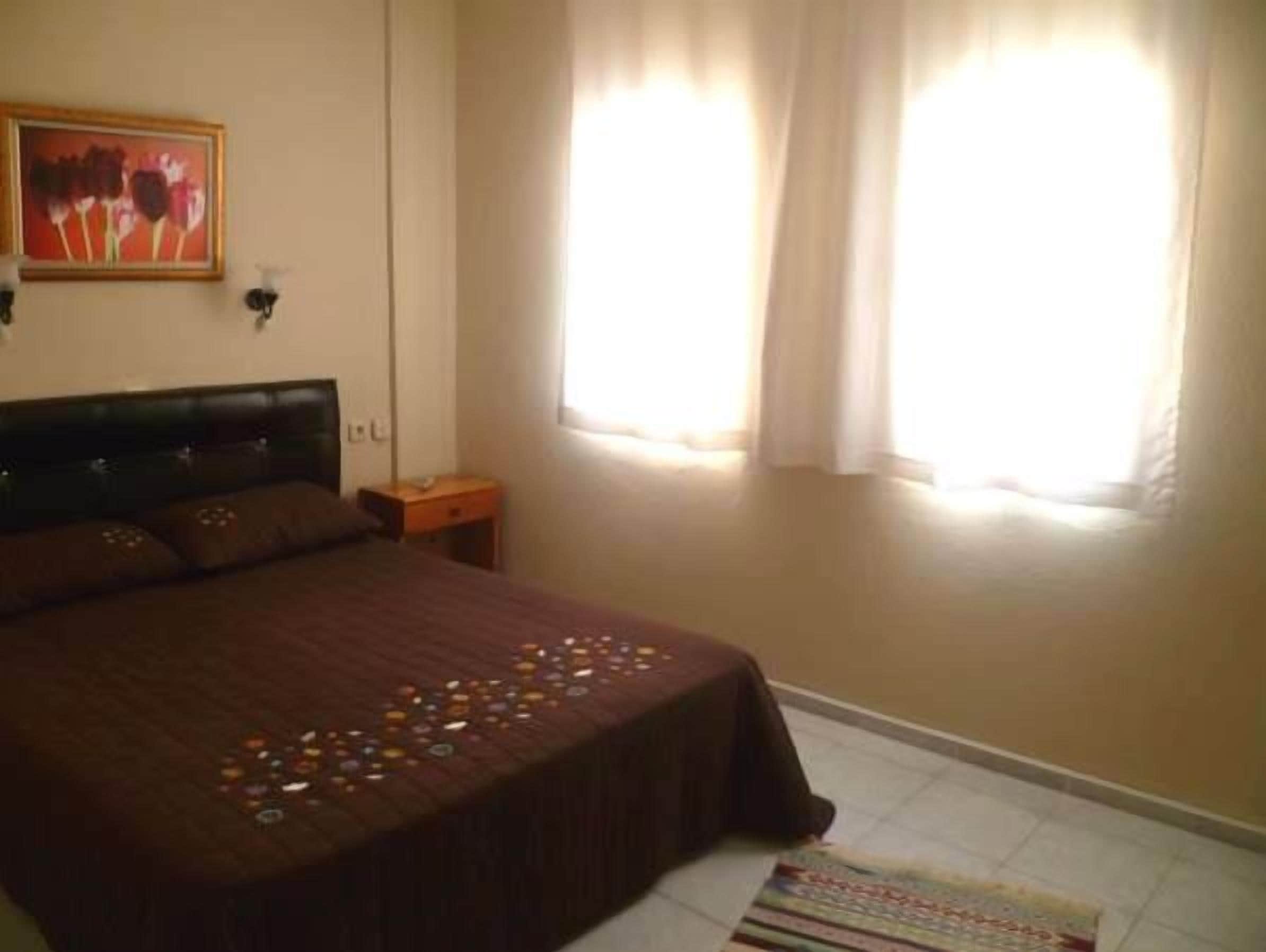 Un dormitorio con cama, mesita de noche, ventana con cortinas y un cuadro en la pared.