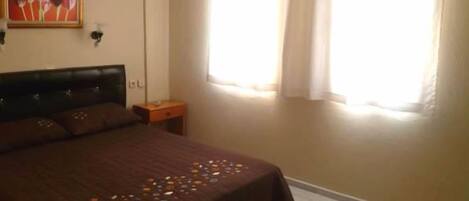 Un dormitorio con cama, mesita de noche, ventana con cortinas y un cuadro en la pared.