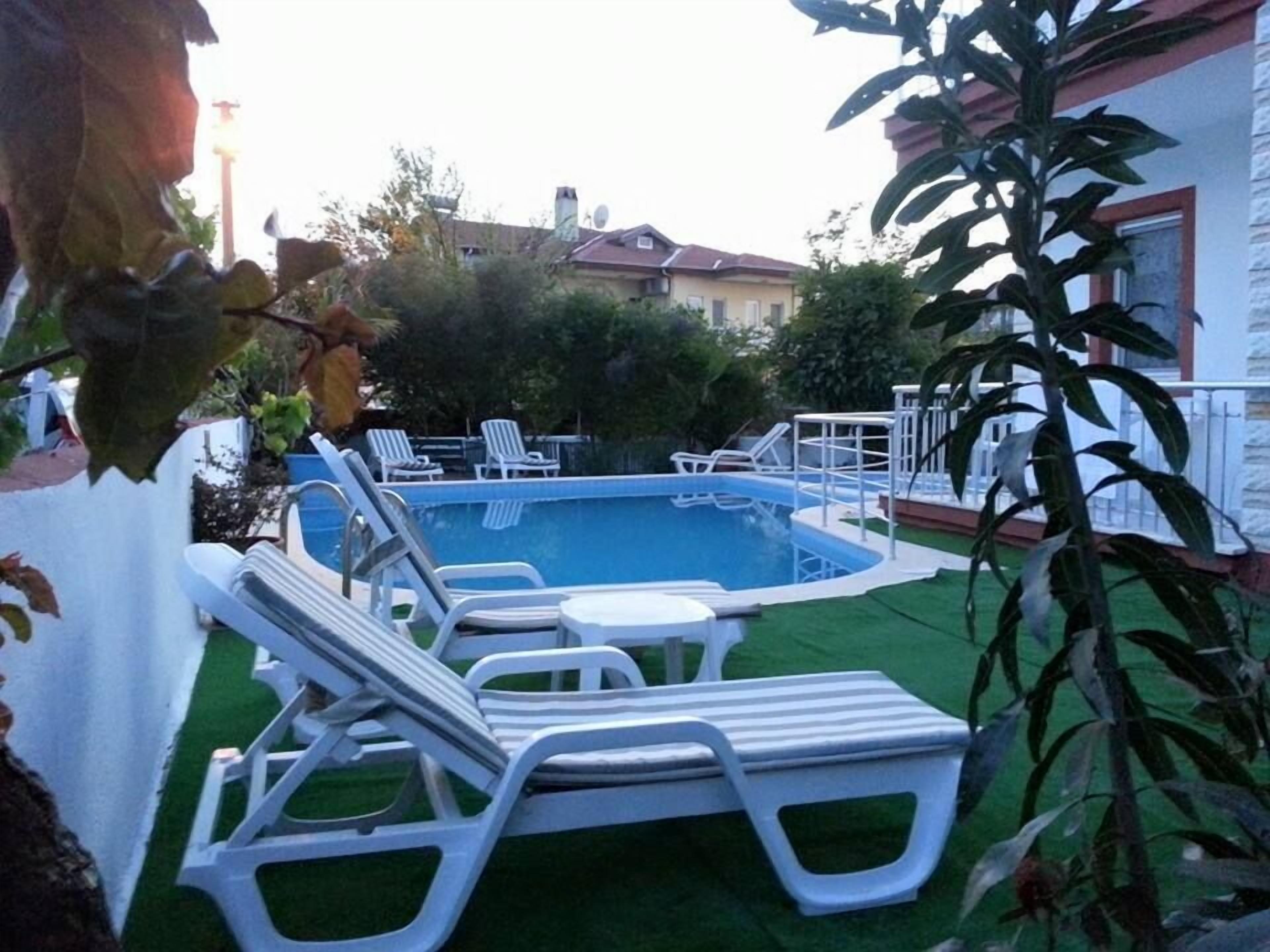 Une piscine entourée de chaises longues et d’une petite table, le tout bordé de verdure et adossé à un bâtiment blanc.