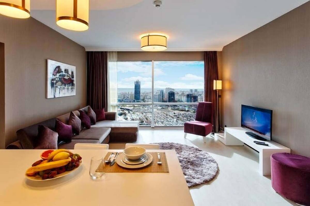 A modern hotel room with a city view, a dining area, and a seating area with a sofa and armchairs.