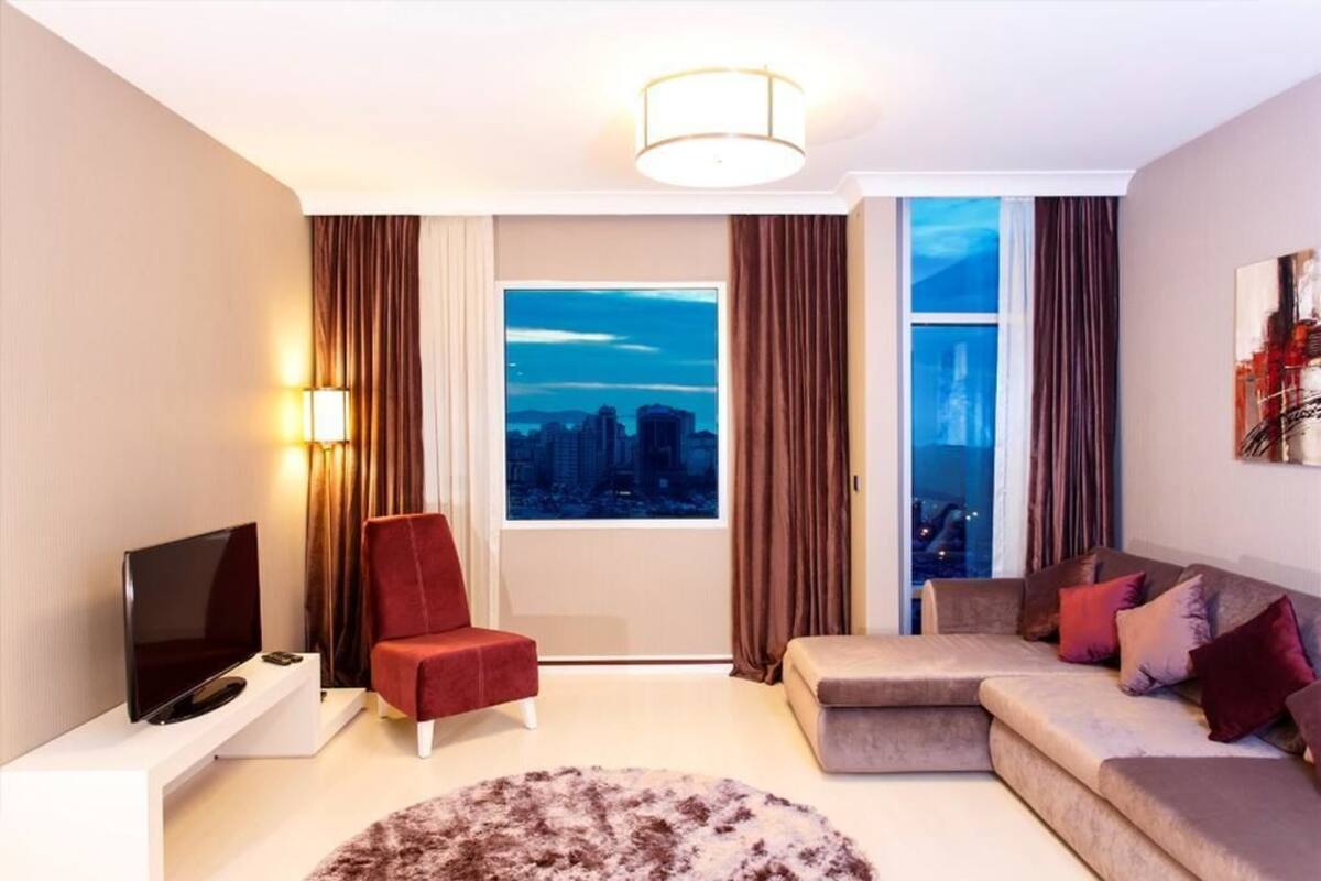 A modern living room with a flat-screen TV, a red armchair, a beige sofa, and a large window with a city view.