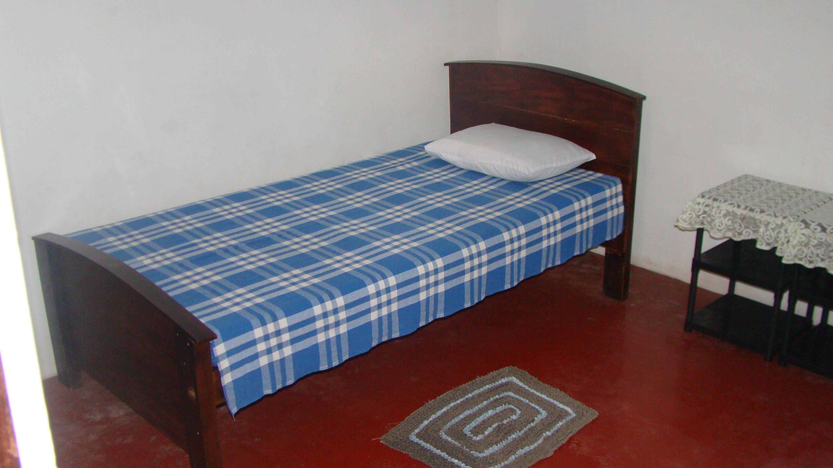 A small, single-bed room with a wooden headboard, a blue plaid bedspread, a white pillow, a small table with a lace tablecloth, and a red carpet.