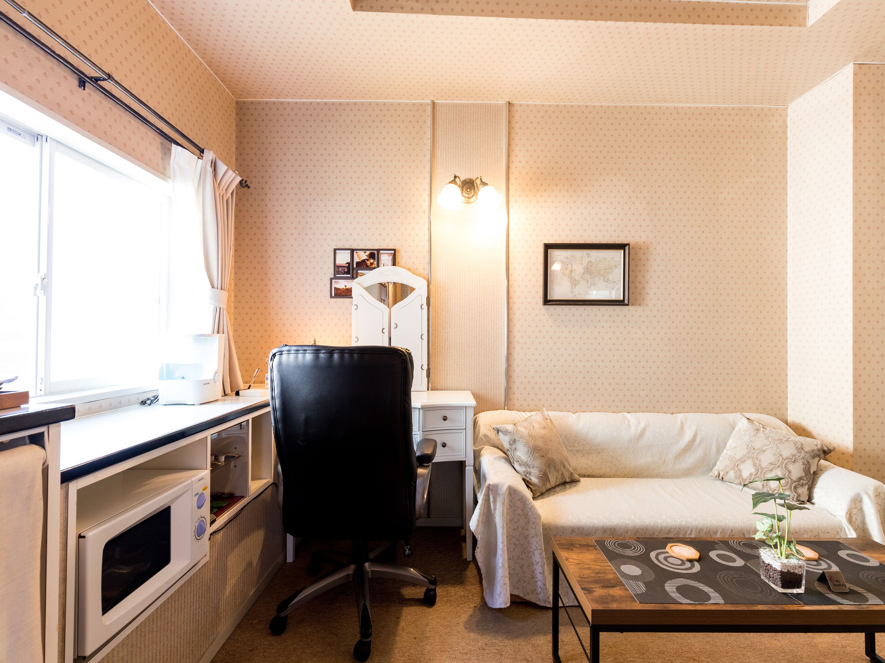 Habitación de hotel compacta con kitchenette, escritorio, silla, cama y una pequeña mesa de centro con una planta.