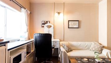 Habitación de hotel compacta con kitchenette, escritorio, silla, cama y una pequeña mesa de centro con una planta.