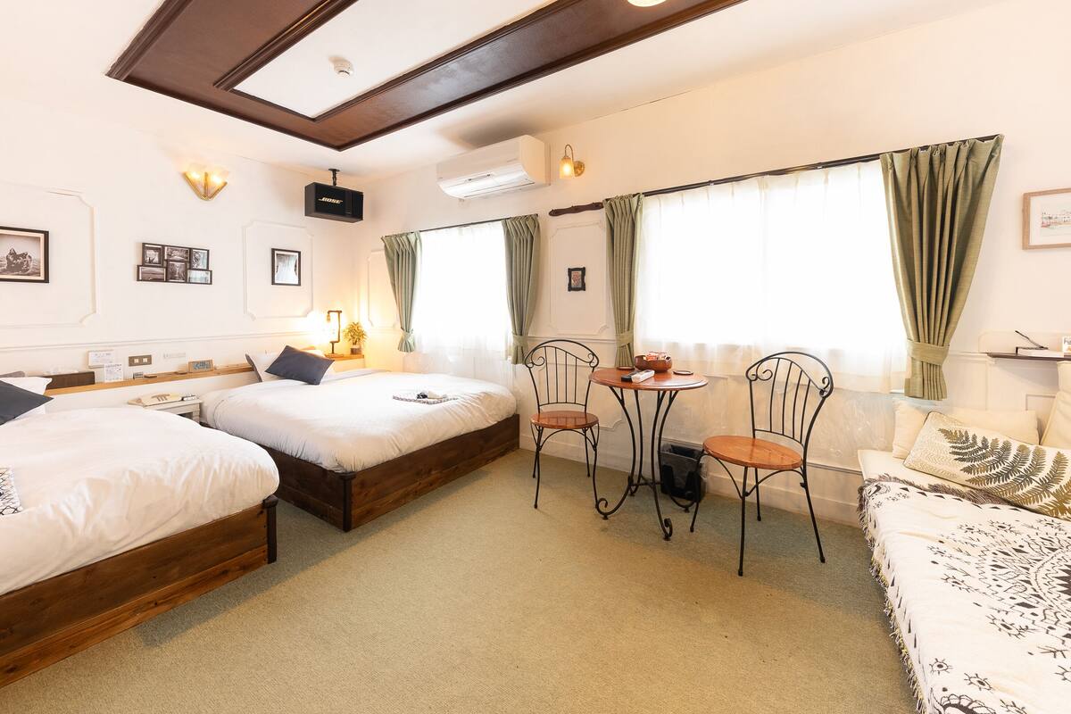 Een hotelkamer met twee bedden, een kleine tafel met stoelen, een bank en een raam met gordijnen.