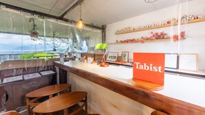 Een restaurantinterieur met een houten toonbank, twee houten tafels, een 'Tabist' bord en een uitzicht op groen door het raam.