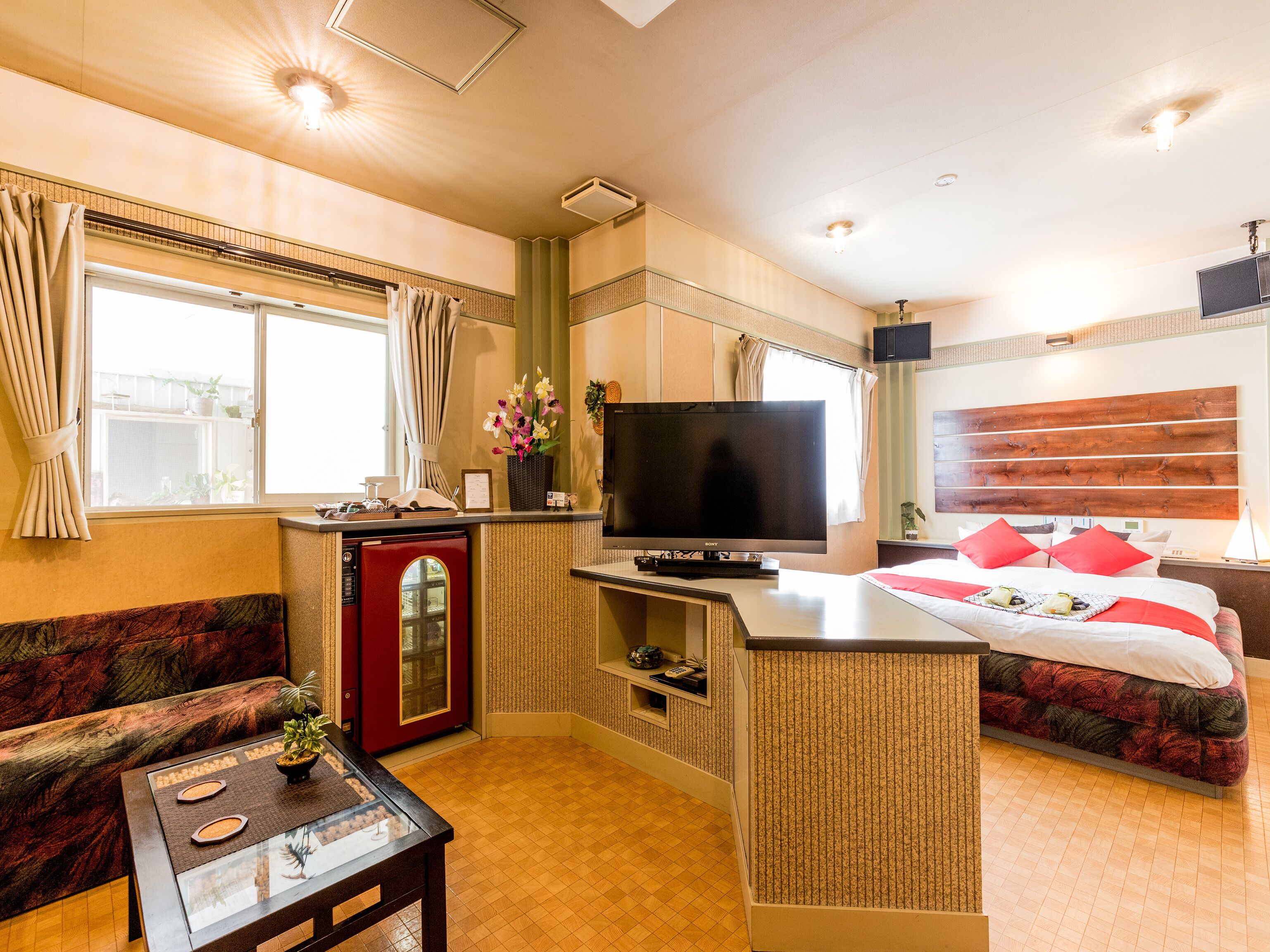 Quarto de hotel compacto com cozinha compacta, TV de tela plana, cama com almofadas vermelhas e sofá.