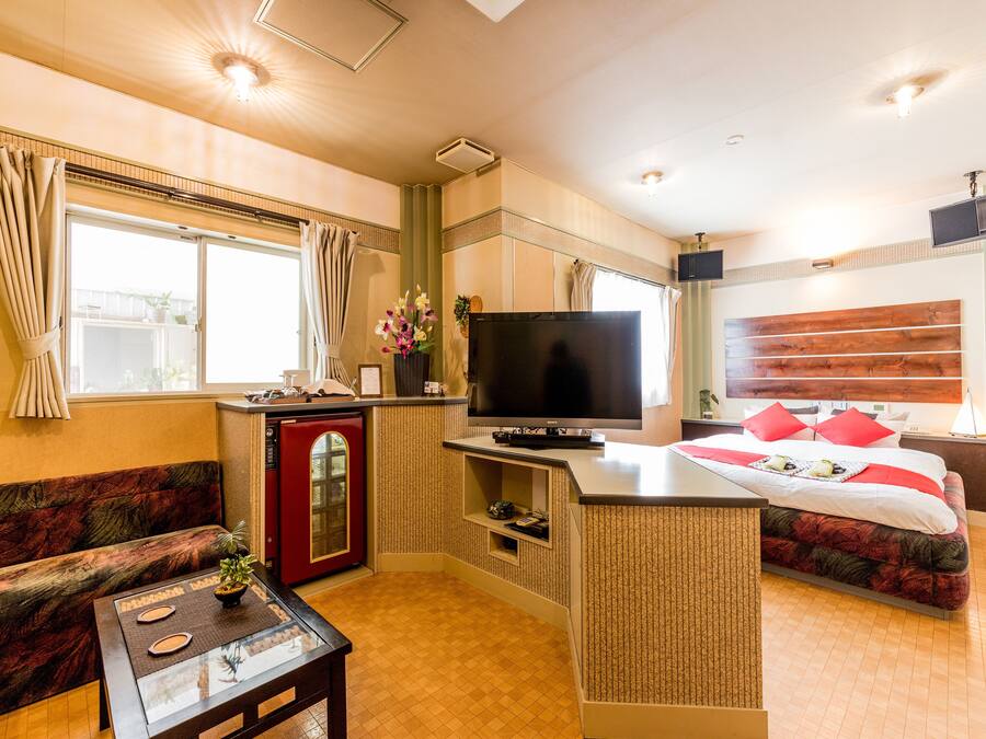 Quarto de hotel compacto com cozinha compacta, TV de tela plana, cama com almofadas vermelhas e sofá.