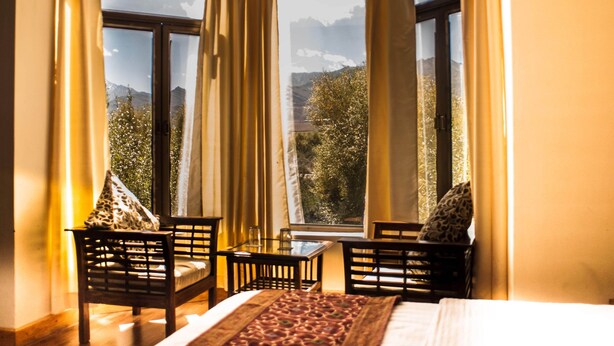 A room with a bed, two chairs, a small table, and a view of mountains through large windows. - Hotel Shivalik - Best Himalaya View Hotel In Almora' (Almora)