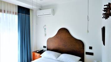 Una habitación de hotel con una cama, una mesita de noche de madera, un teléfono, un aire acondicionado instalado en la pared y un ventilador de techo.