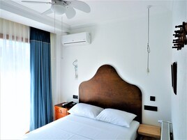 Una habitación de hotel con una cama, una mesita de noche de madera, un teléfono, un aire acondicionado instalado en la pared y un ventilador de techo.