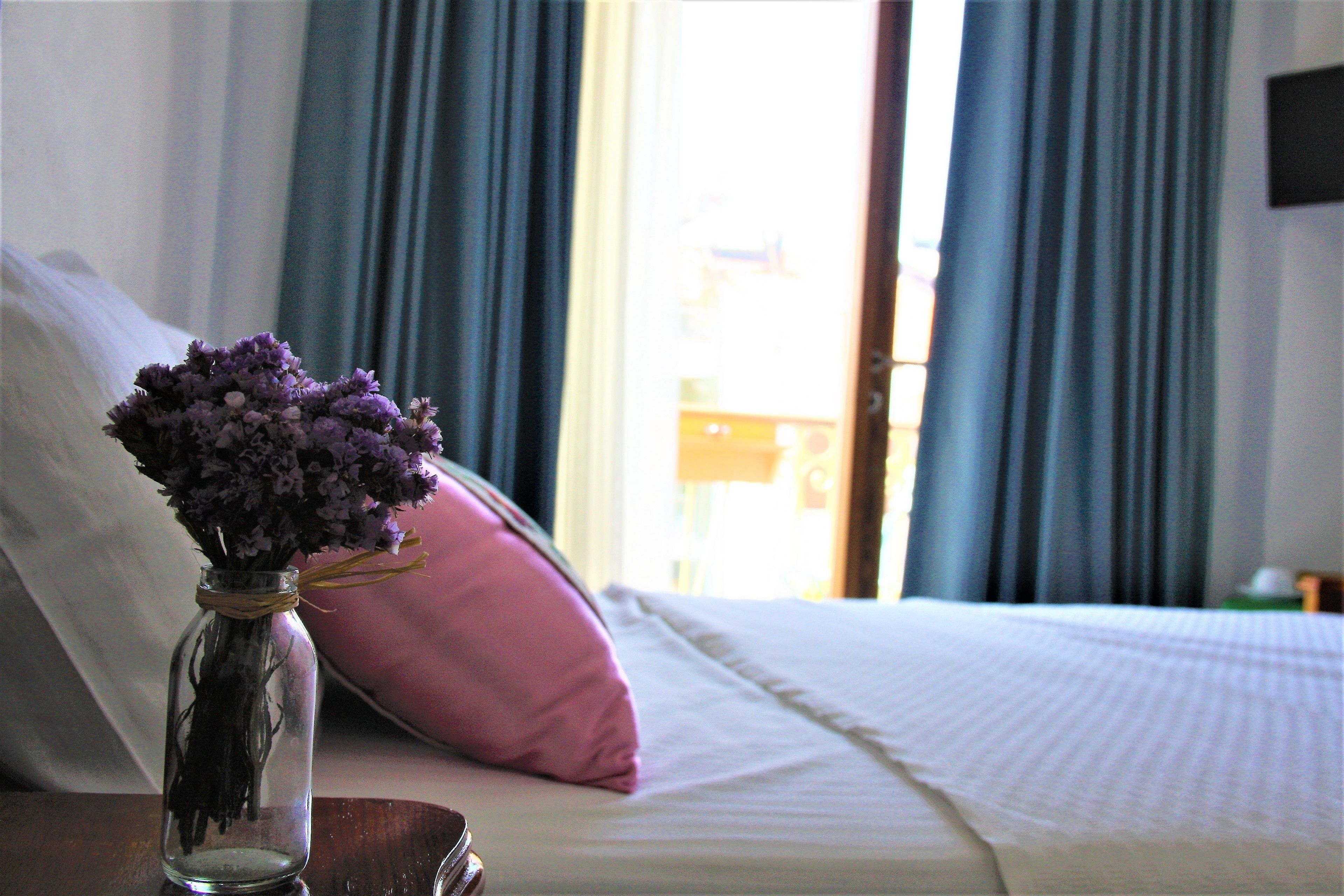 A bed with a pink pillow and a clear glass vase with purple flowers on a wooden nightstand.