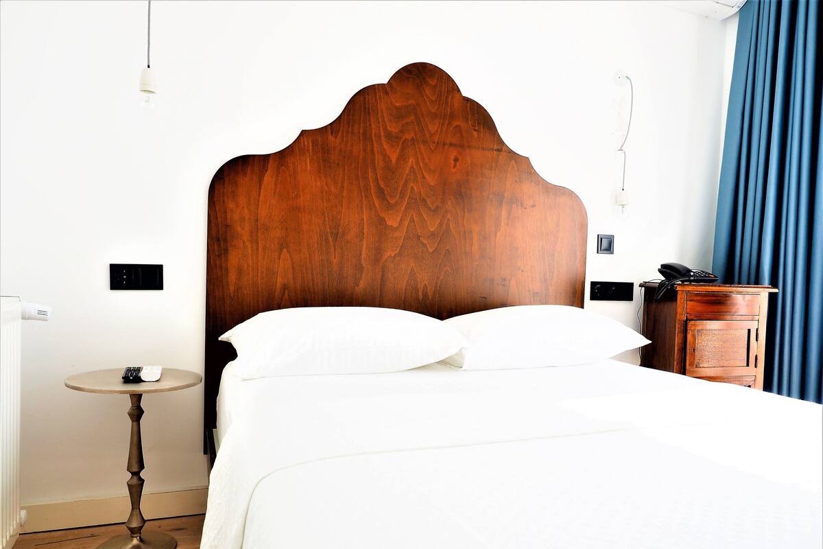 Un lit bien fait, avec du linge de lit blanc, une tête de lit en bois, une table de chevet avec une télécommande et une petite armoire contenant un téléphone.