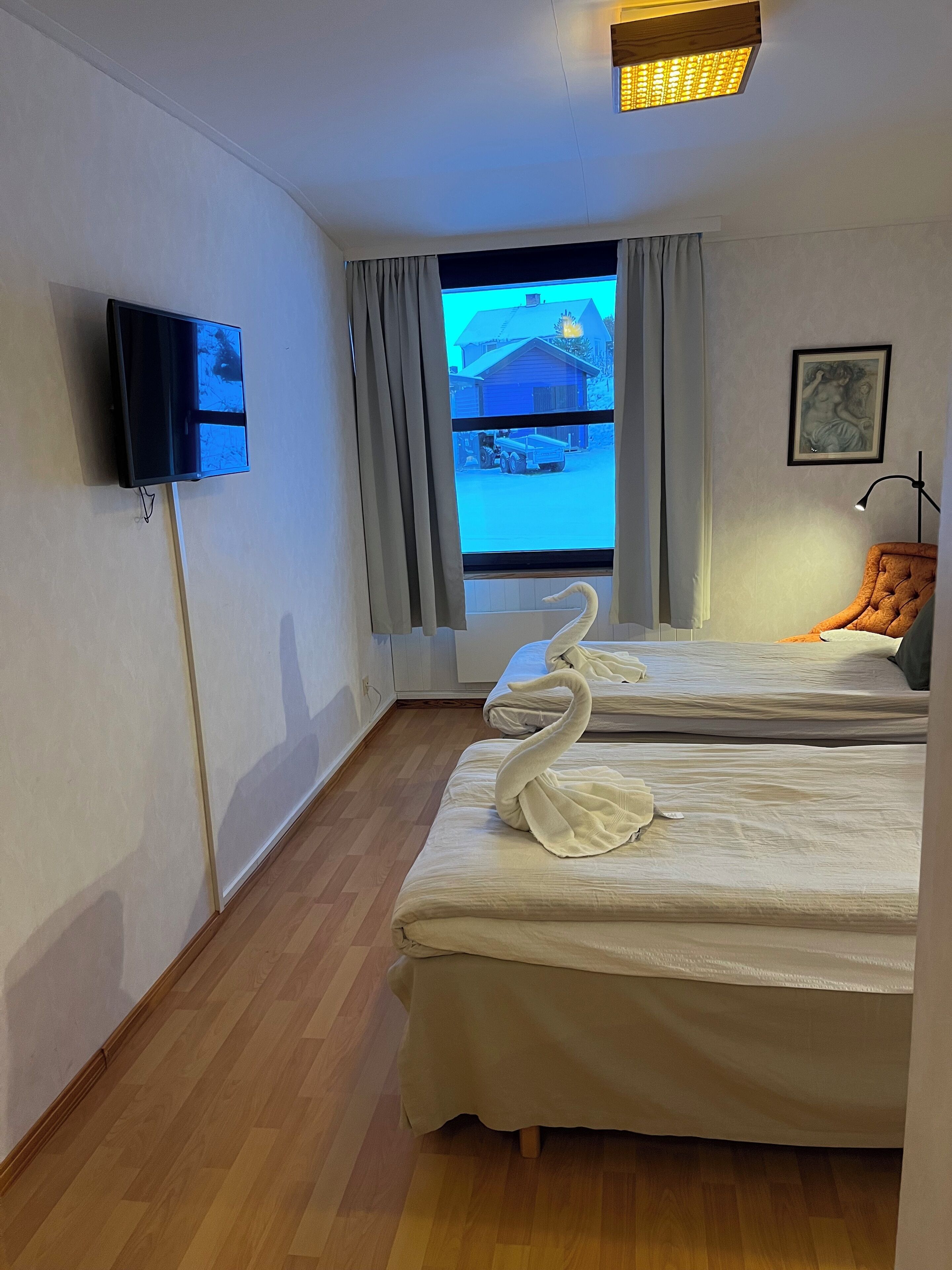 A hotel room with two beds, a TV mounted on the wall, a window with curtains, a framed picture on the wall, and a lamp on a side table.