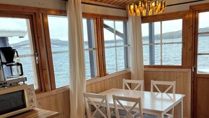A wooden interior with a dining table and chairs, a coffee machine, and large windows overlooking a body of water.