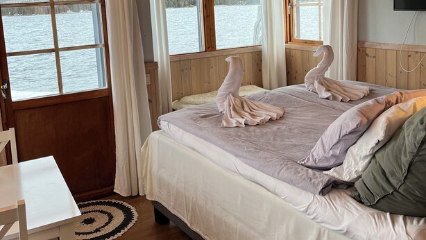 A bedroom with a wooden ceiling, a bed with a white and gray duvet, two swan-shaped pillows, a white chair, and a view of a lake through a window.