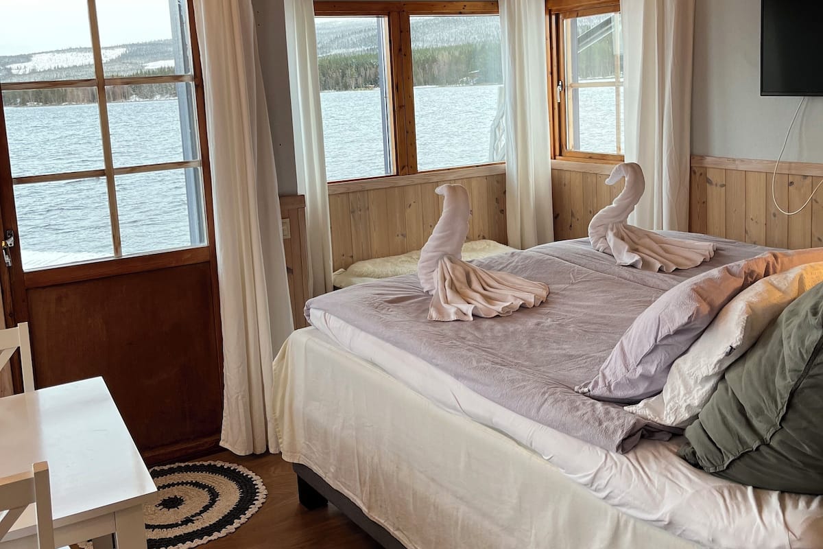 Une chambre avec un plafond en bois, un lit recouvert d’une couette blanche et grise, deux oreillers en forme de cygne, une chaise blanche et une vue sur un lac par la fenêtre.