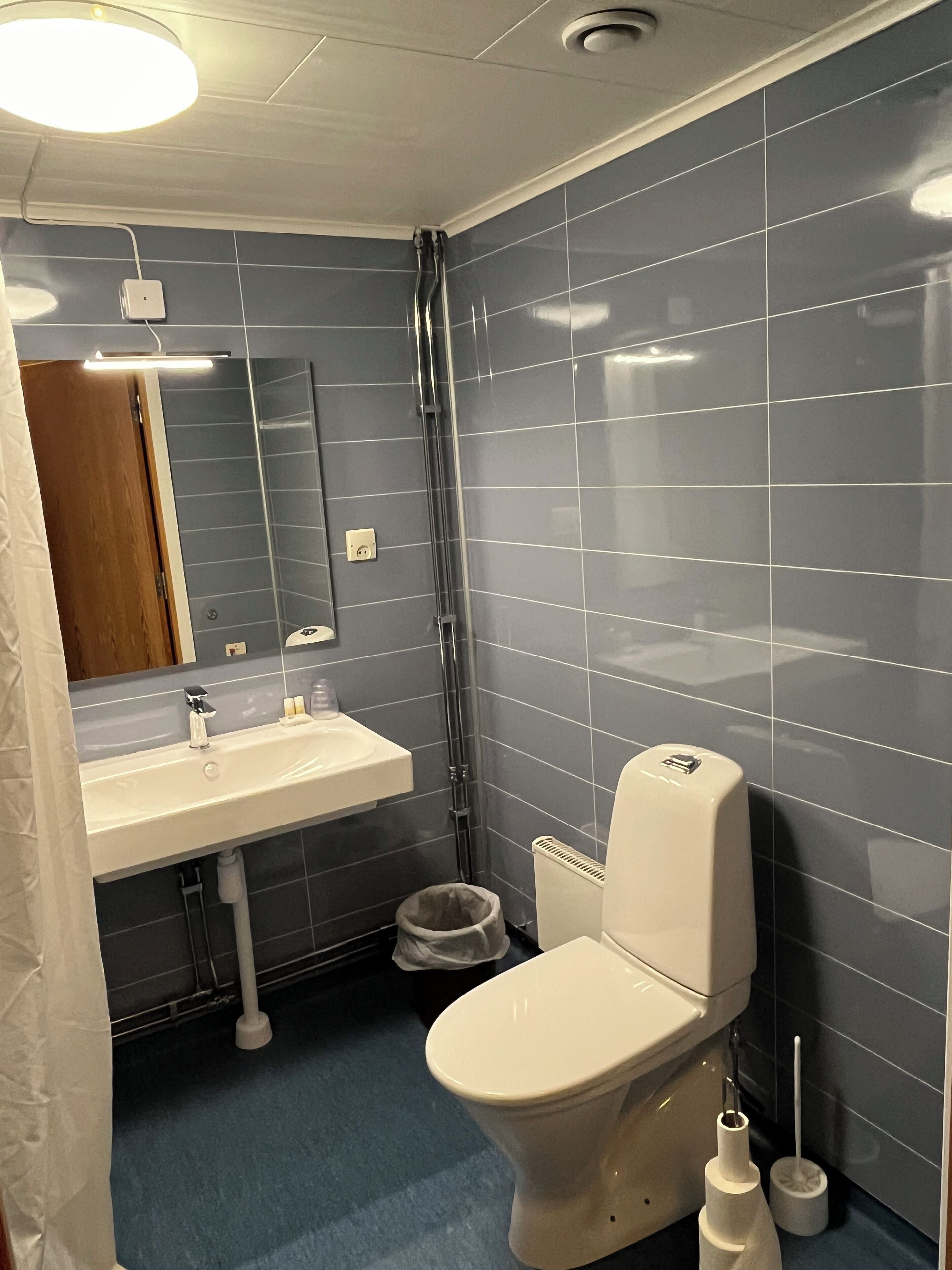 Un baño con un lavamanos blanco, un espejo y un inodoro. Las paredes están revestidas de azulejos oscuros y el suelo es azul.