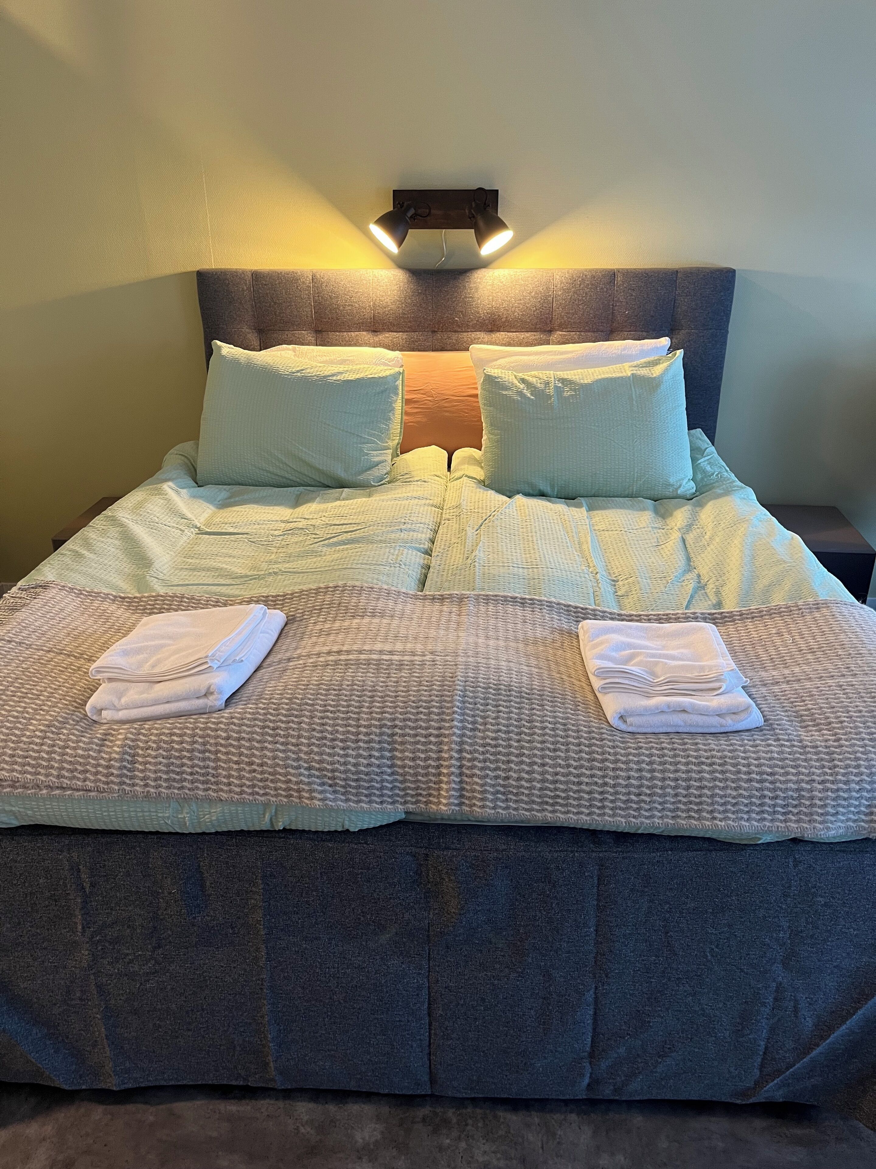 Un lit bien fait, avec une tête de lit, deux oreillers et des serviettes pliées, posés sur une couverture à motifs.