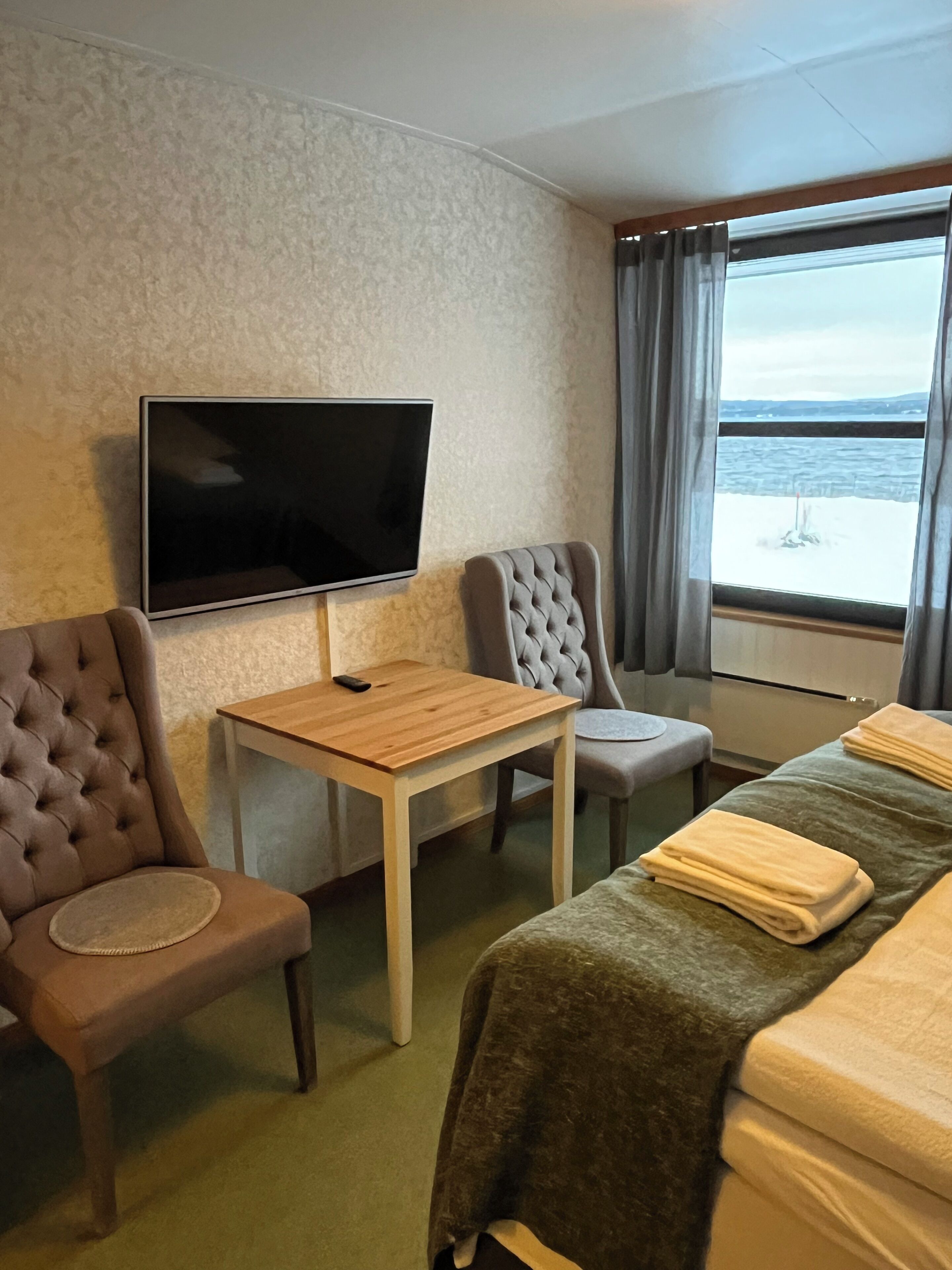 A hotel room with a bed, a TV mounted on the wall, a small table, two chairs, and a window with a view of the sea.