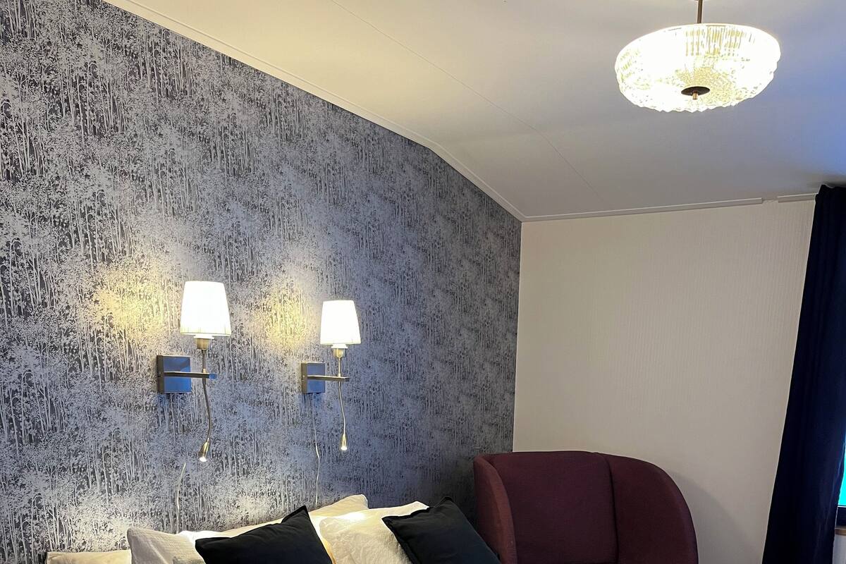 Un lit bien fait, avec des oreillers blancs et noirs, un plaid vert et deux lampes fixées au mur.