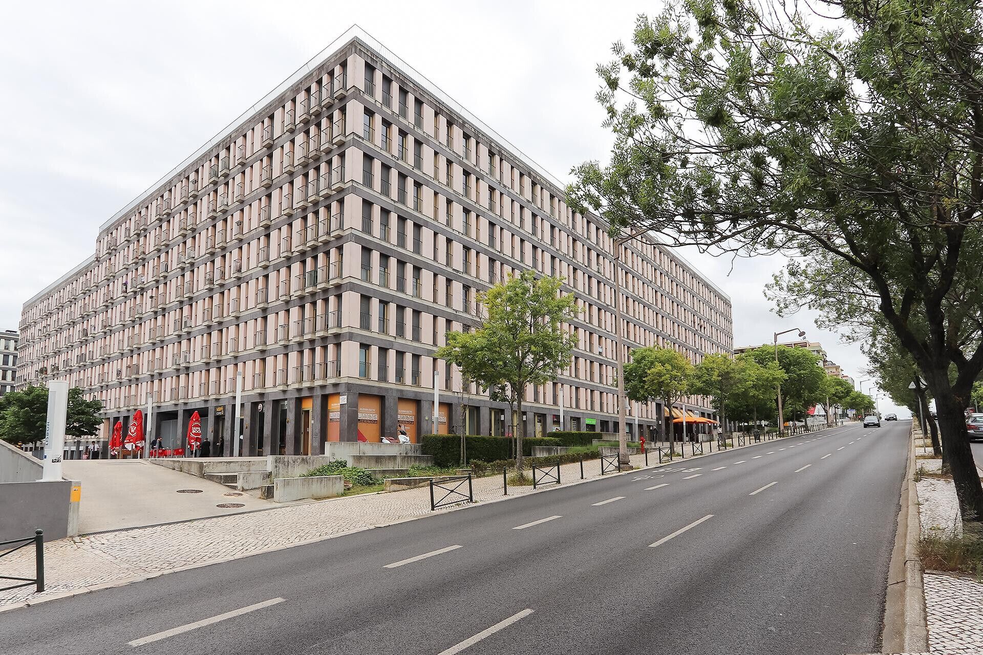 A modern multi-story building with a grid-like facade, a paved street in front, and trees lining the sidewalk.