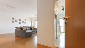 A spacious living room with a grey sofa, wooden flooring, and a door leading to a kitchen area. - Modern & Bright by Homing (Lisbon)
