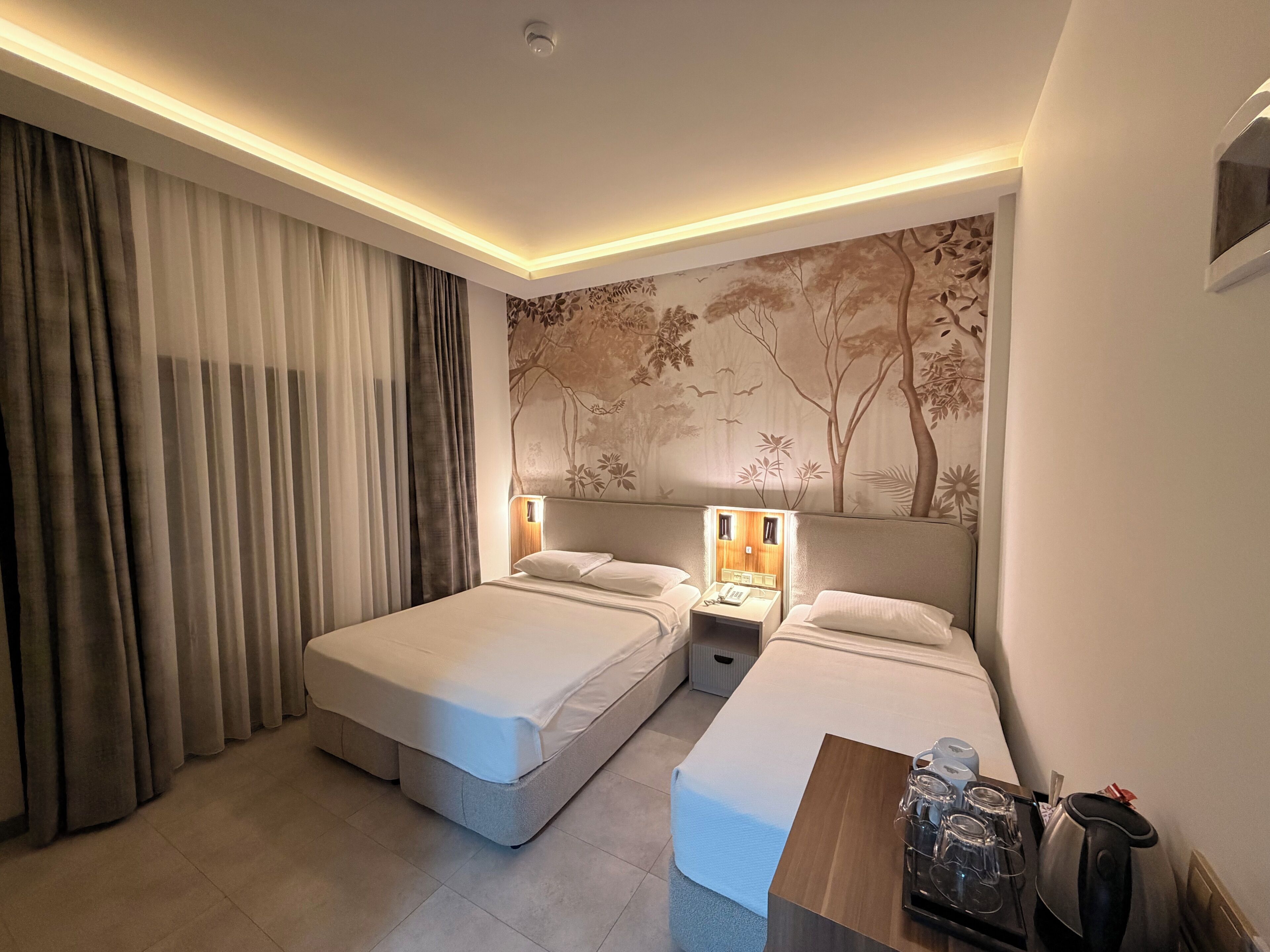 A hotel room with two beds, a nightstand with a lamp, a wooden side table with glasses and a kettle, and a large tree mural on the wall.