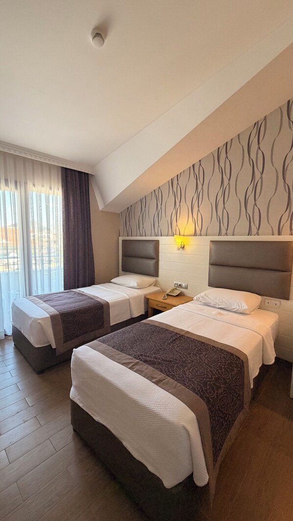 Habitación de hotel con dos camas, una mesita de noche con lámpara, una ventana con cortinas y un papel tapiz estampado.